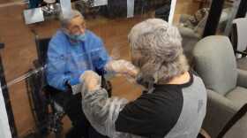 Two women wear gloves and hold hands through holes cut in a clear plastic sheet.