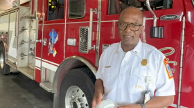A uniformed fire department officer stands in front of a red fire truck holding a circular, white smoke alarm.