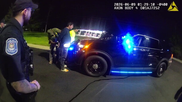 Two men are being handcuffed by a border patrol agent as they stand in front of the hood of a police car. In the foreground, a police officer with a Southgate police emblem on his shoulder watches the scene. It is dark, but a brightly-lit gas station can be partly seen in the background. 