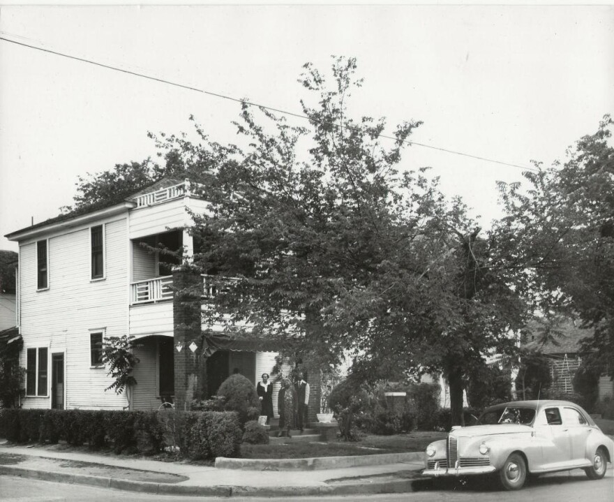 A black and white photo of a large white corner house. A car is parked outside and a family stands near the front door.