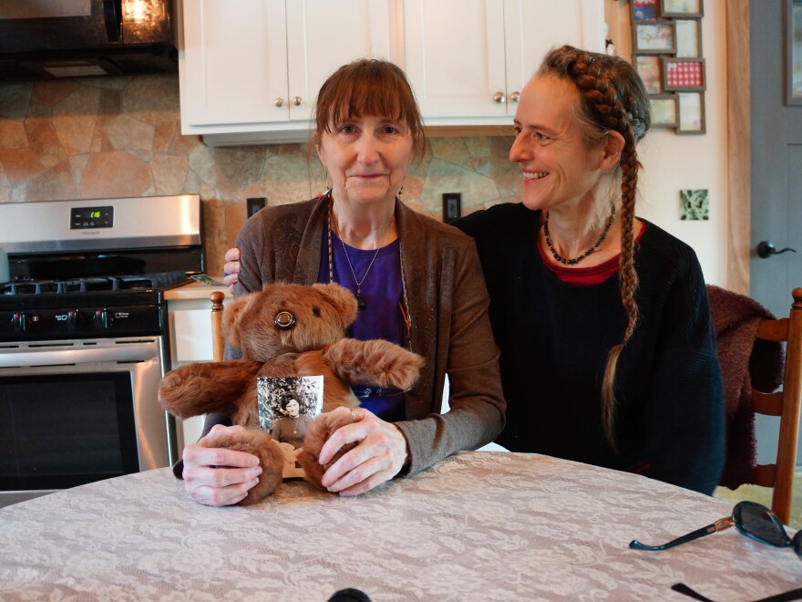 Two women sit at a kitchen table with a teddy bear, smiling