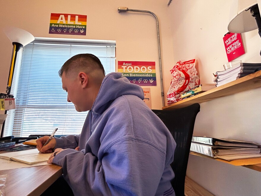 A person with short hair and wearing a gray hooded sweatshirt sits at a desk and writes in a book.