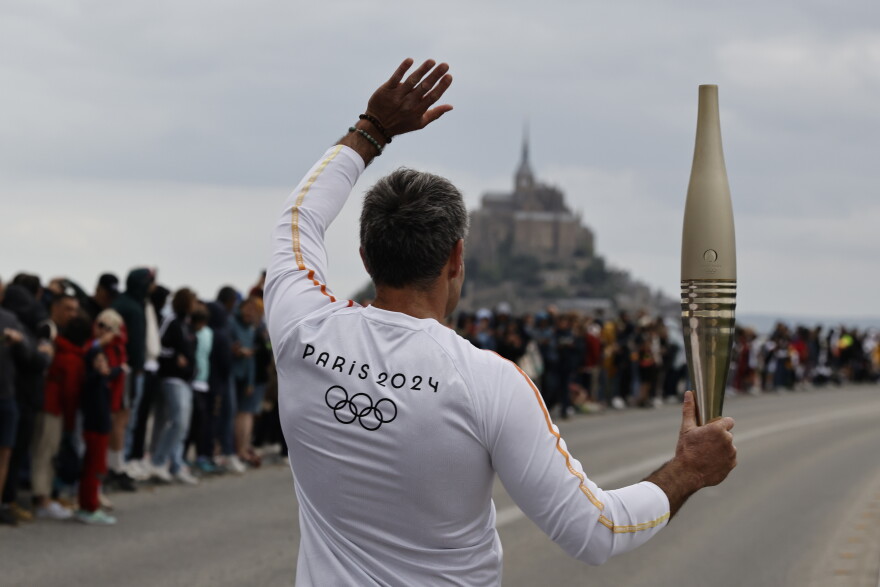 Torchbearer Maxime Gohier waves to the audience as he holds the Olympic torch in front of the Mont-Saint-Michel, Friday, May 31, 2024 in Brittany. (AP Photo/Jeremias Gonzalez)