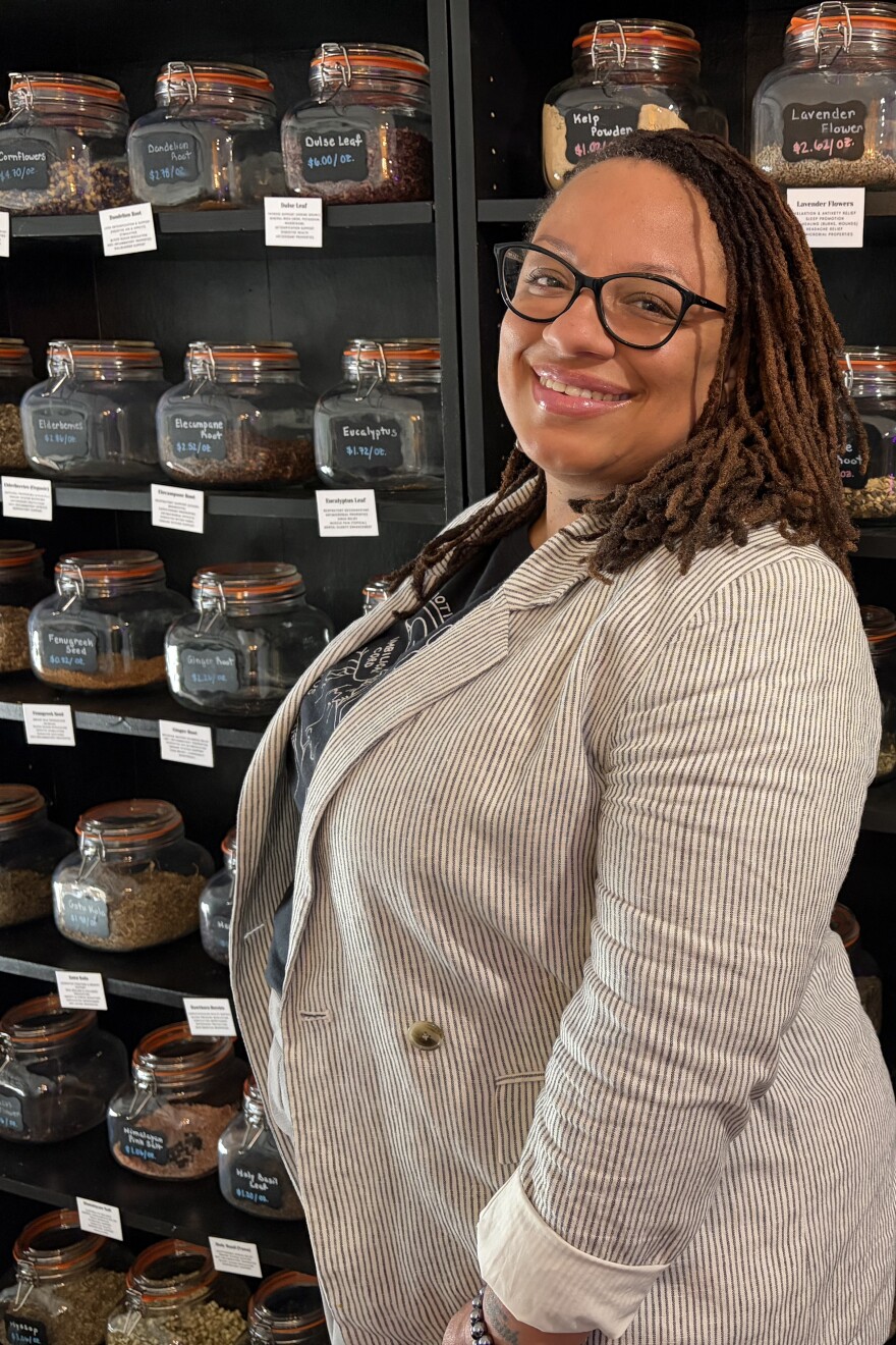 Erica Dickson stands to the right side of the image in front of a large black shelving unit with bulk herbs in large mason jars. Each jar has a blackboard label with the herb's name and price. Below each jar is a white card displaying information about the herb's medicinal uses. Dickson wears a white and gray pinstripe blazer over a black shirt with a white lineart diagram of a baby in a womb. Dickson is looking slightly over her shoulder and smiling at the camera.