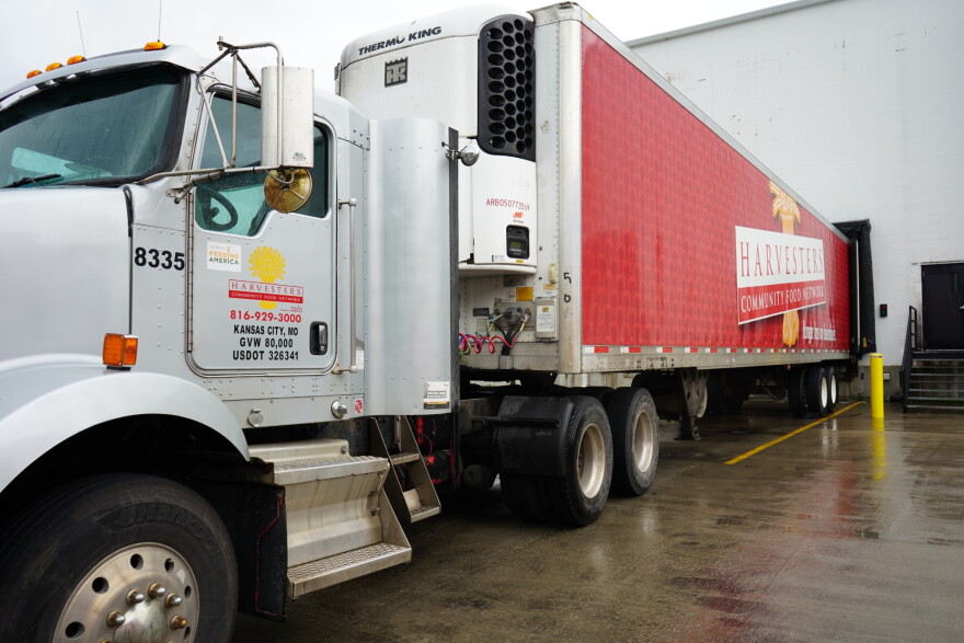 A large semi truck is parked at a warehouse. The trailer is completely red with a large Harvesters logo pasted in the middle of it's face.