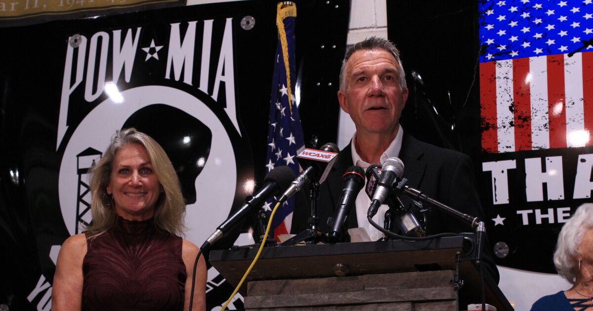Photo: vermontpublic.org A man in a black jacket and white button-down shirt stands at a podium, speaking into mics. A woman with blonde hair stands