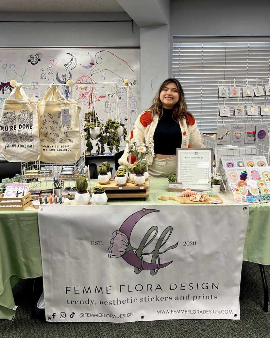 Christine Almendras poses behind her table for her business. On the table are different items for sale and in the from is a banner that reads, Femme Flora Designs.