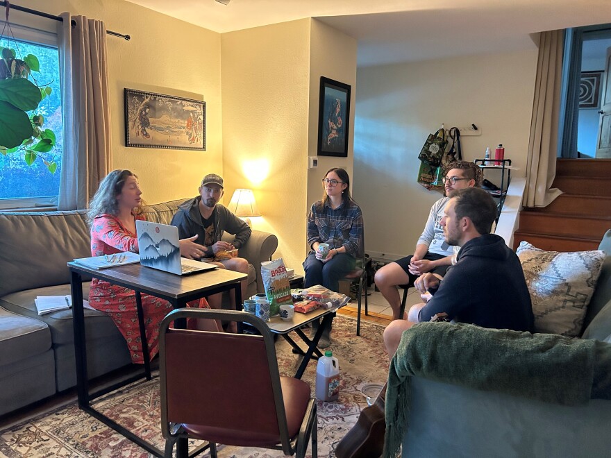 A group of young adults sit on couches and chairs in a circle inside a cozy home and talk with one another. 