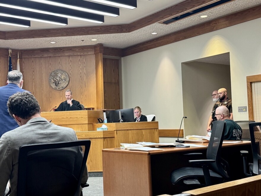 A courtroom scene with a judge at the bench, a court clerk near a computer, a lawyer speaking, people seated at tables, and a person in handcuffs escorted by an officer near a side entrance.