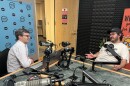 Matthew Peddie on the left wears a social white shirt, black glasses and watch. Bryce Bonsack on the right, wears a white shirt with a black hat. They are at a radio studio, with microphones in front of them. 