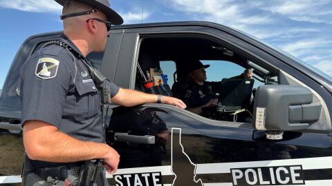 An Idaho State Police trooper stands next to an ISP vehicle, seemingly talking to the driver. 