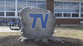 A large rock with the letters TV painted in blue with a gold outline placed next to a school building.