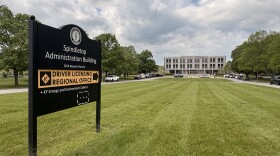 A sign in the foreground on a long, green lawn leading to a three-story building in the background under a cloudy sky. The sign reads "SPINDLETOP ADMINISTRATION BUILDING 2624 RESEARCH PARK DR. DRIVER LICENSING REGIONAL OFFICE"