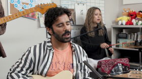 Close-up of Rafay Rashid playing guitar and singing. He's wearing a black and white striped shirt over a peach t-shirt. Talia Temporal plays a drums in the background.