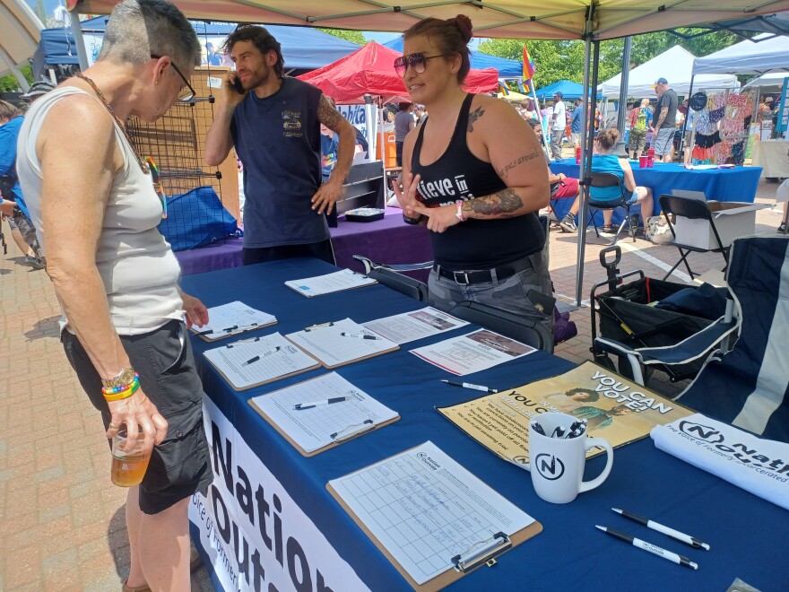  The Nation Outside information booth at Kalamazoo's Pride Festival on June 3, 2023.  Petitions and literature sit on top of a blue tablecloth.  There's a mug with pens in it. Tenee Gildea is on one side in a black tank top talking to someone about the issues the group supports, like Michigan's Fair Chance Access to Housing Act.  