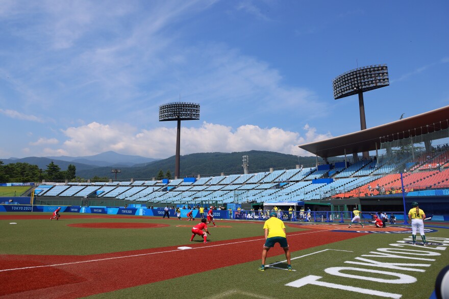 Softball players wearing red and yellow uniforms stand play a game on the pitch, with rows of empty seats and mountains in the backdrop.