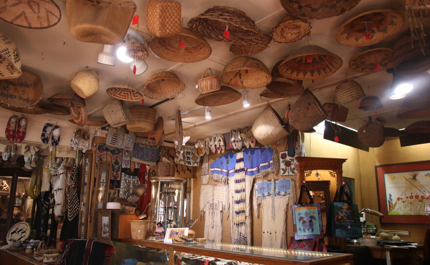Some water baskets hang from the ceiling. Every inch of wall space is covered with paintings, clothing, moccasins, and jewelry.