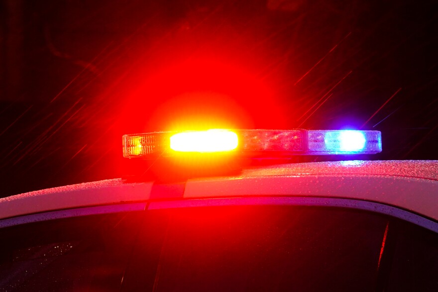 Shown are the red and blue flashing lights of a police vehicle, zoomed in to show only the lights and the top half of the police cruiser. Rain is trickling down through the frame, illuminated by the flashing lights. 