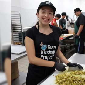 A smiling young woman with a baseball cap and apron wears black food-service gloves as she chops a pile of something on a white cutting board. Behind her more young adults work in a commercial kitchen filled with stainless steel counters and pans. 