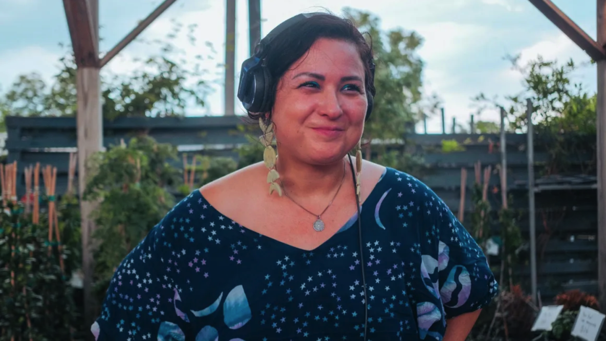 Midshot of Bonnie Ilza Cisneros. She is smiling with her head turning 3/4 to her left side. Her hands are on either side of her hips and dressing a beautiful dark turquoise with stars and petal-like figures.