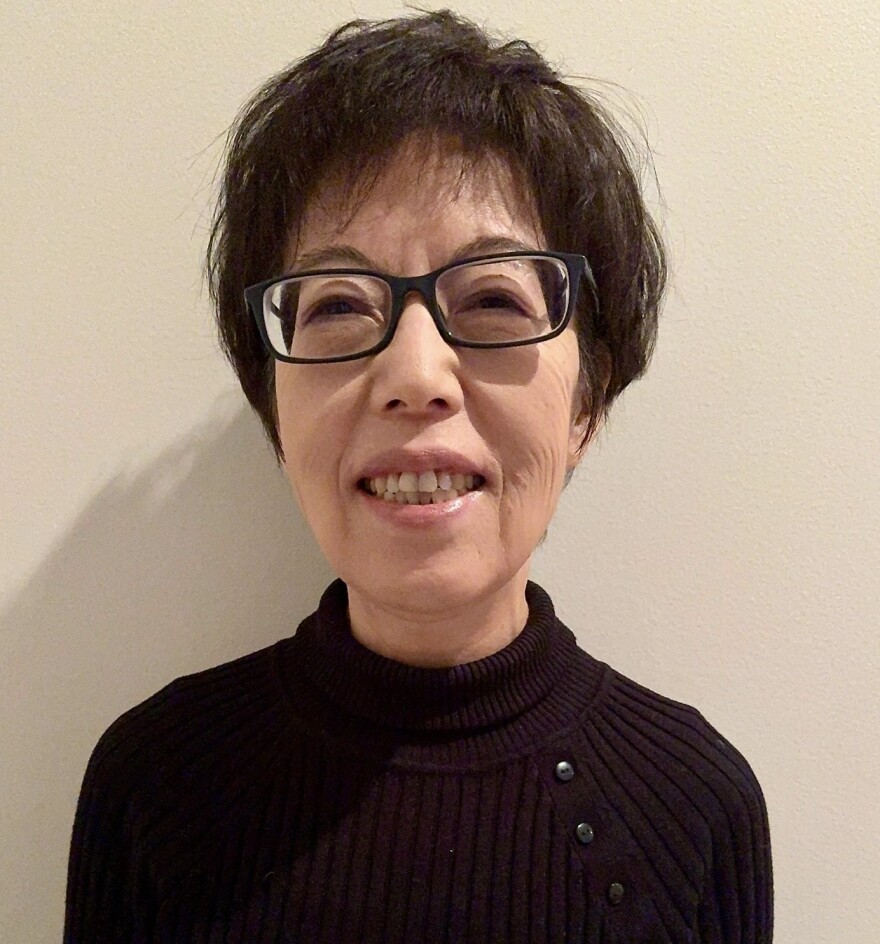 A photo of an Asian woman smiling against a white wall. She has short black hair and glasses. She's wearing a black sweater.