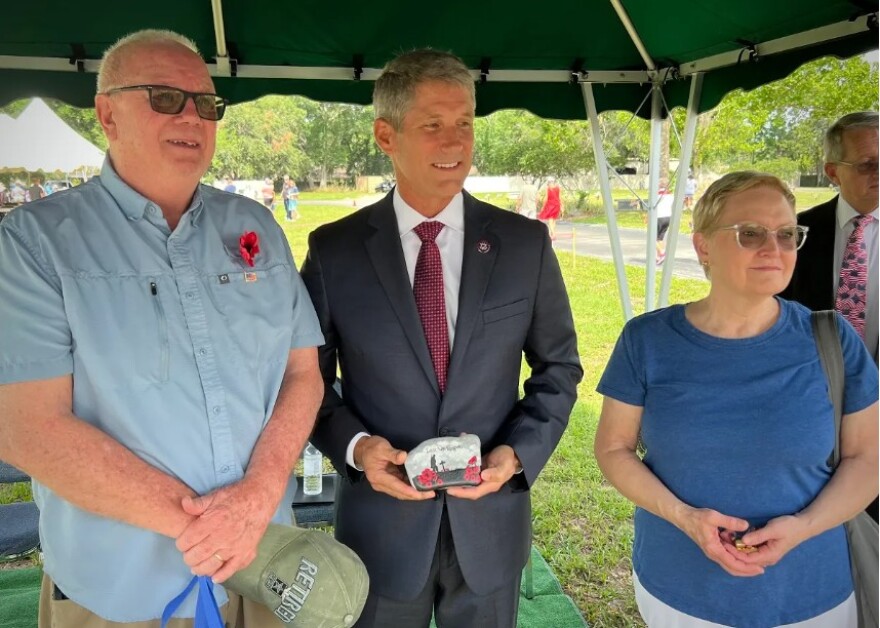 Man in a gray suit  and red tie flanked by an older man with sunglasses in a blue short and a woman with glasses and blue blouse