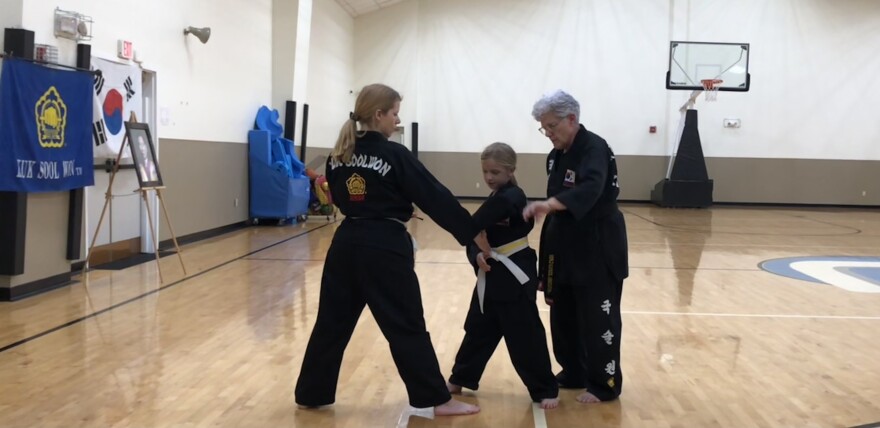 Two women and girl wearing black martial arts robes practice in a gymnasium with a wooden floor and white walls.