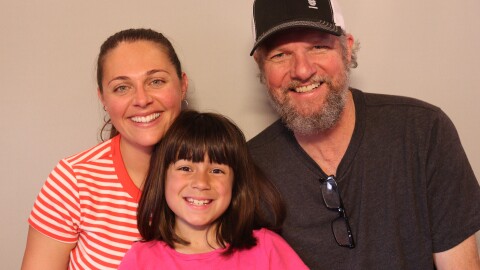Taylor and Ronin Salazar and Mike Rothwell at their St. George StoryCorps appointment in May 2025.  Mike and Taylor stand side by side with Ronin in front between them. All of them smile with teeth. Ronin is a young girl of 8 years old. She has brown eyes and shoulder length black-brown hair with bangs to her eyebrows. She wears an orange tee shirt.  Taylor rests her chin on Ronin's head. She wears an orange and white striped tee with orange ribbing around the neckline.  She has green eyes and wears four small silver hoop earrings. Mike's hair, visible beneath his black and white baseball cap, is gray.  He has a full, trim gray beard.  Mike's eyes are green and he wears a dark gray tee shirt with black frame glasses hanging from the neckline.