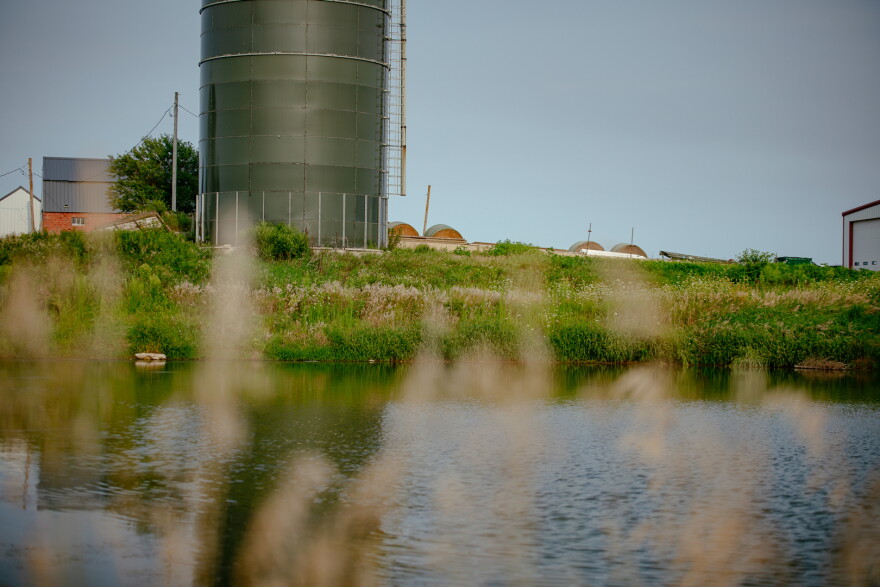 Across a pond and up a bank of grass, sits a grain elevator, barn and hay bales.