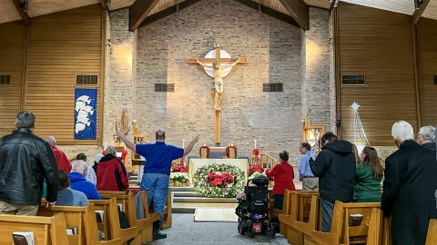 Congregants stand and sit during worship as part of a inclusive Christmas Mass at St. Bernadette Catholic Church. There is a wheelchair user in the isle near the front, facing an altar decorated with Christmas wreaths and lights.