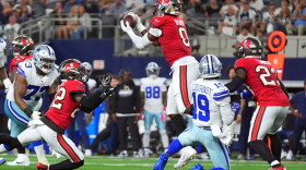 football action with player #0 in red jersey and pewter helmet and white pants jumping to catch the ball. teammates are on each side along with opposing players in white jerseys and gray pants and helmet