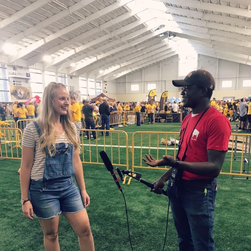 Blonde woman in overalls talks to a reporter in a red shirt and baseball hat, holding a microphone, at a football game