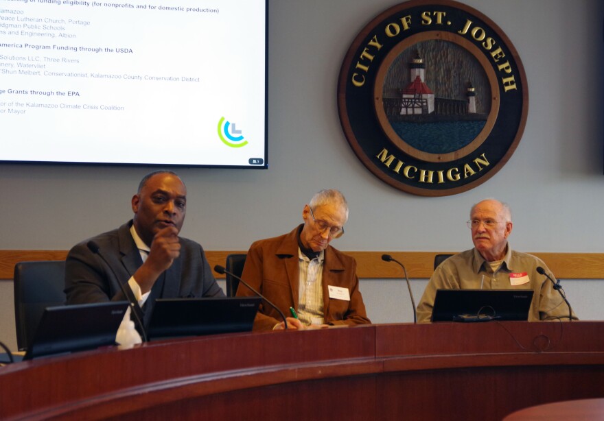 Three well dressed men sit at a round table, the seal of the City of St. Joseph sits on the wall behind them. 