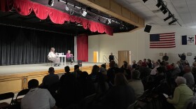 Two women sit on a stage in front of a large crowd in an auditorium