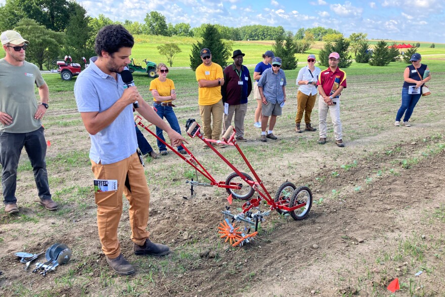A man with a microphone points to tools with one and two wheels in a field with small leafy greens. A crowd stands around him.