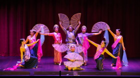 A group of eight dancers pose on stage. Four hold wide fans while the others hold blue or pink scarves.