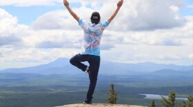 Person wearing tie-dye Tshirt standing atop a mountain with leg up in yoga position and arms outstretched