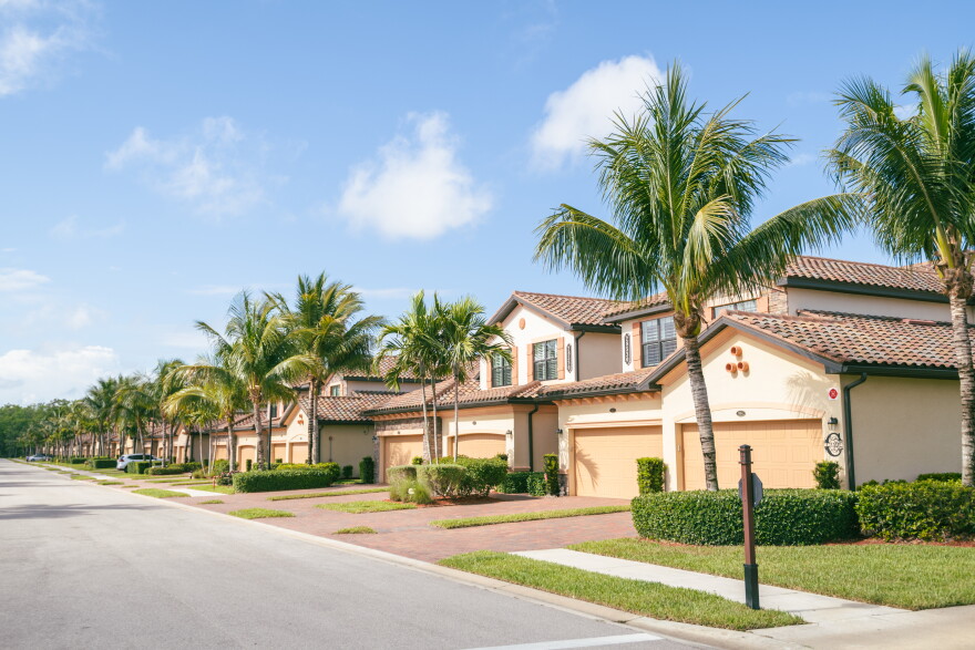 A row of side by side, tan two story houses with big garage doors in a Florida neighborhood with palm trees and bushes in front of the houses.  