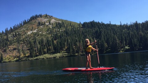 A child wearing a yellow life jacket paddleboards on a calm lake with forested mountains rising behind her under a clear blue sky.