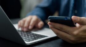 Mans hands working on laptop while holding a cell phone.