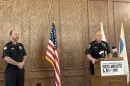 Two police officers stand indoors near an American flag. One speaks at a podium with microphones and a sign reading "Choose Greater Peoria Red, White & Boom!" while the other stands to the side, listening.