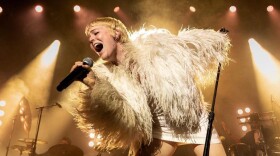 A woman in a shaggy top leans toward the audience while singing into a microphone on stage at a concert.