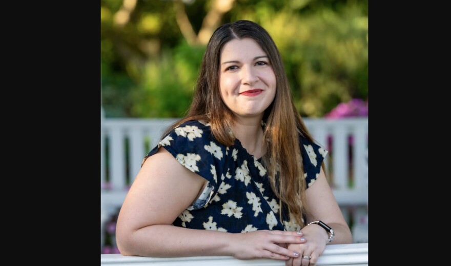 A woman in a blue dress with a flower pattern leans on a railing as she poses for a portrait photo with trees in the background. 