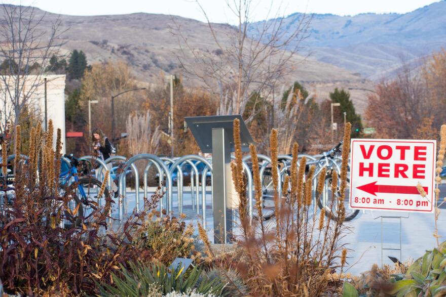 A red and white "Vote Here" sign with bike racks and mountains in the background. 