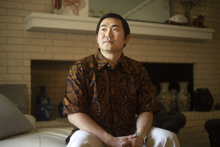 Ronald Muljadi sits with his hands folded sitting in a living room with a brick fireplace behind him.