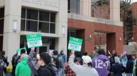 A crowd carrying signs stops at the front of a large brick building, with 'HOVEY HALL' denoting the building name.