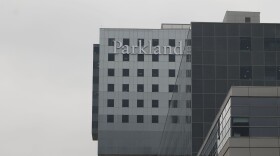 Parkland Hospital on a very overcast day. One top of a dark glass structure is a gray structure with a sign that reads "Parkland" in the upper left corner. 