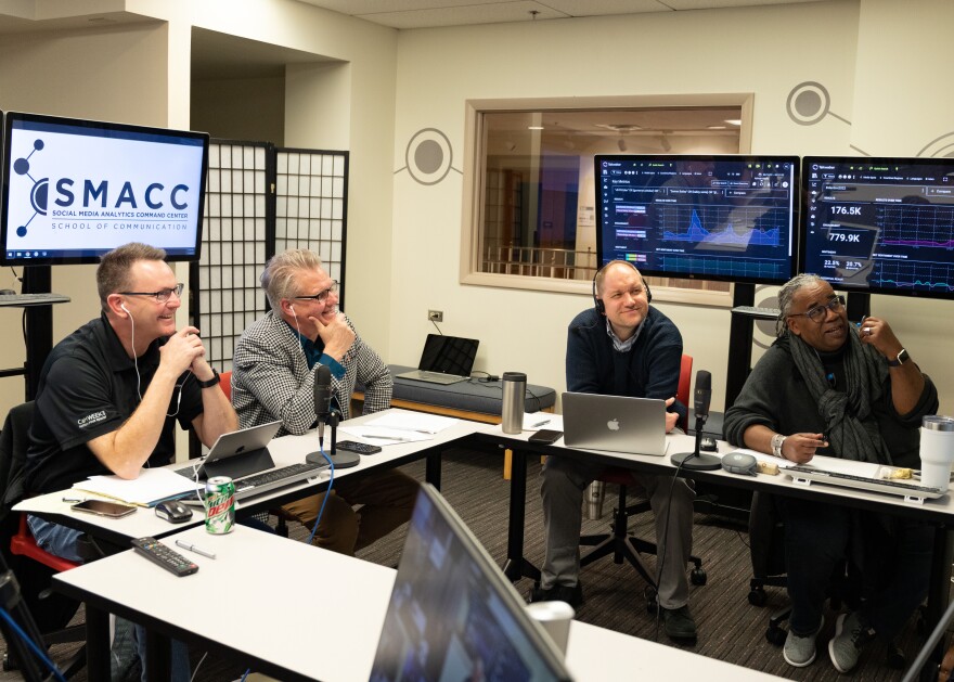 Four men are seated chatting at two long tables positioned at 90-degrees. They smile and look at television screens out of view that are broadcasting a video call.