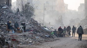 People sit around a fire and walk past collapsed buildings on February 13, 2023 in Hatay, Turkey.