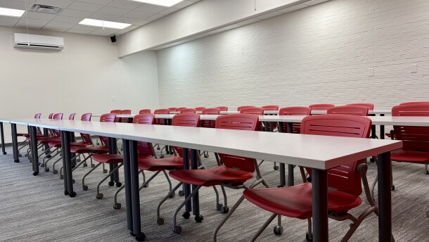 Three rows of red plastic office chairs sit at three long empty tables in a new classroom. 